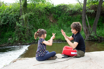 Two children, a teenager and a girl, playing children's games at a camp. High-fiving while singing, sitting on the ground next to a waterfall of a river that runs through a forest. 