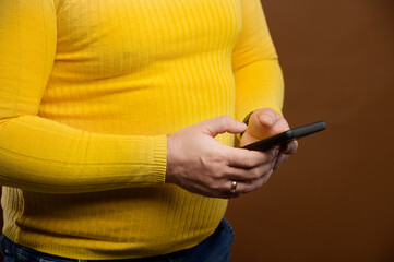 Close-up of a plump middle-aged man in a yellow casual jacket uses his phone while typing or surfing the Internet. Studio shot on brown background