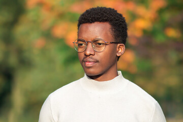 Naklejka na ściany i meble Portrait of sad serious upset pensive thoughtful black guy, young African Afro American man in glasses walking in a sunny golden autumn park at fall, thinking. Natural background.