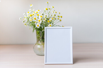 Portrait white picture frame mockup on wooden table. Glass vase with camomile
