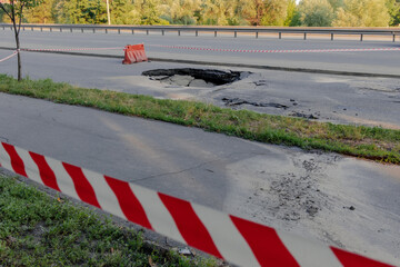A hole in the ground in the middle of the road surrounded by a red tape. A large pit in the asphalt due to a pipeline accident