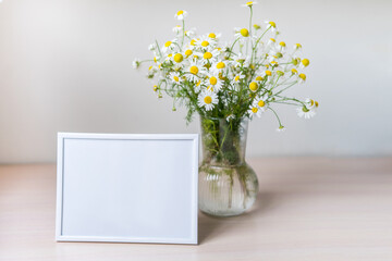 Portrait white picture frame mockup on wooden table. Glass vase with camomile