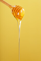 Honey drips in a thin stream from a honey dipper on a yellow background.