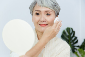 Senior Asia woman is holding a mirror and using puff to make up herself.