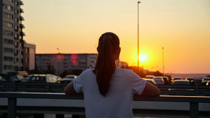 unrecognizable teenage girl with long hair in ponytail admires setting sunset in orange sky standing on overpass bridge backside upper view