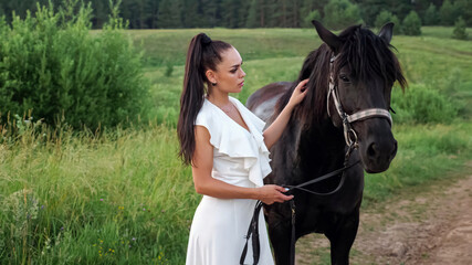 Young woman brunette with long hair in ponytail pets black horse and animal shakes head standing on...