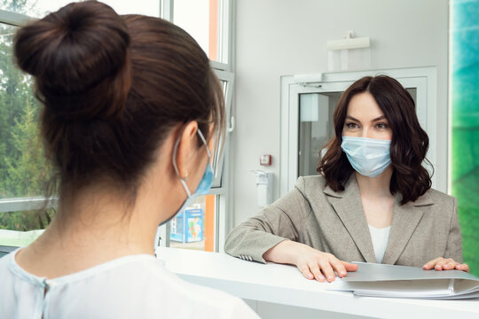 Pretty Brunette Young Woman Patient With Protective Mask Talks To Female Administrator Leaning On Counter At Reception In Contemporary Hospital Close View