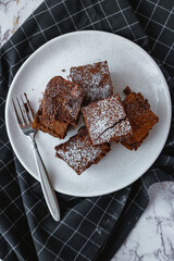 Brownie squares dusted with powdered sugar with a fork on a white plate and black kitchen towel. Flatlay.
