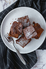 Brownie squares dusted with powdered sugar with a fork on a white plate and black kitchen towel. Flatlay.