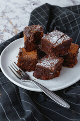 Stacked brownie squares dusted with powdered sugar with a fork on a white plate and black kitchen towel. 