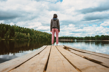 Fototapeta na wymiar The view from below from the wooden pier of the forest lake to the girl standing alone with her back