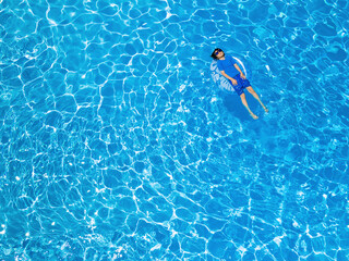 Aerial drone view of  a young boy of ten years old wearing sun protection t-shirt and sunglasses is lying on the inflatable ring in the swimming pool. Summer holidays concept, copy space on the left.