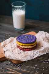 Stacked, colored cookies on a piece of baking parchment and small cutting board with a glass of milk. Wooden background.  Scattered lavender. 
