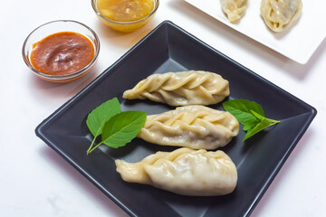 Traditional chicken meat stuffed steamed momos or dumplings in a black plate along with soup and tomato sauce on white background.