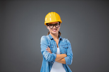 A woman in the business world. Portrait of a young female engineer standing in front of a gray background with her arms crossed. The girl is casually dressed with a protective helmet on her head