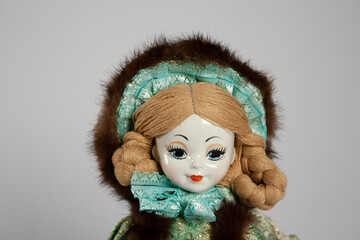 close up of the face of a porcelain doll, with braided hair and a fur cap with green lace trim on a soft gray background with copy space