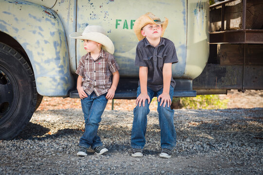 Two Young Boys Wearing Cowboy Hats Leaning Against an Antique Truck in a Rustic Country Setting..