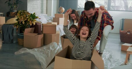 Young fun happy parents push excited cute children in cardboard boxes on the floor moving in new apartment slow motion.