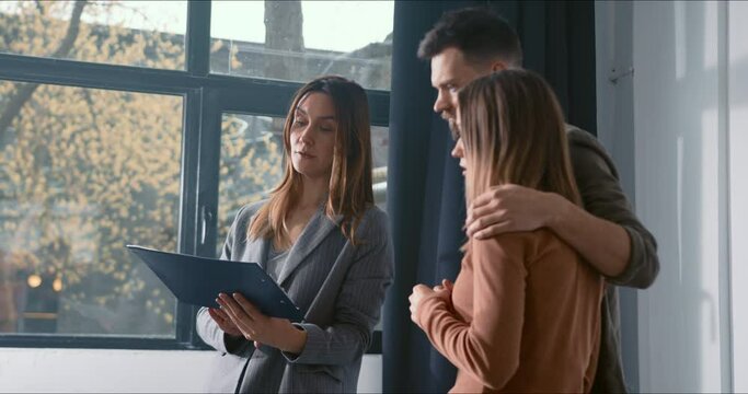 Attractive real estate agent broker woman consults young happy couple showing them new spacious bright apartment on sale