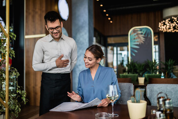 Hansome young waiter showing menu to beautiful female customer in restaurant.