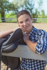 Young girl standing with her hands resting on the wooden fence in the horse ranch. Smiling and posing for the camera. Leather saddle hanging on the wooden fence in the foreground.