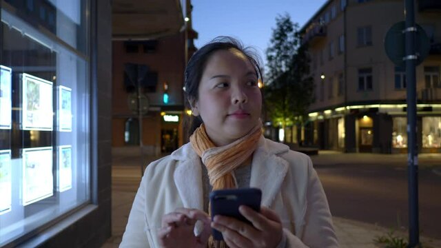Asian woman wearing scarf walking and using smartphone on the footpath in the city. Slide on the screen and texing on phone on the street. Building and moving car background. City night concept