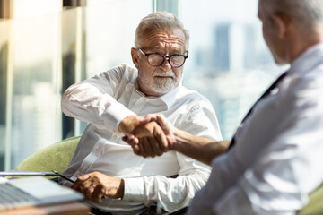 Picture of young business man shaking hand with his older business partner. They are in white shirt and black tie. They are sitting on a table in a hotel lobby. 
