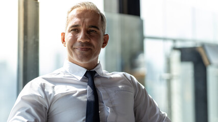 Picture of young business man smiling. He is wearing white shirt and black tie. He is sitting on a table in a hotel lobby.