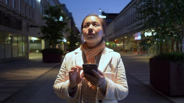 Front view of Asian woman standing, using smartphone and looking around in the city. Slide on the screen and texing on phone on the street. Buildings and shop background. City night concept