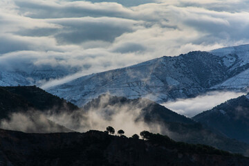 Landscape of mountains covered with clouds, snow and a viewpoint next to some trees in the Sierra Nevada.