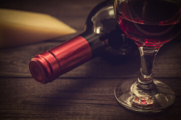 Bottle of red wine with a glass of red wine and a piece of parmesan on an old wooden table. Close up view, focus on the bottle of red wine