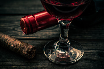 Bottle of red wine with a glass of red wine and cuban cigar on an old wooden table. Close up view, focus on the cuban cigar