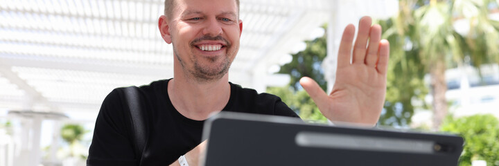 Portrait of smiling man waving to tablet screen while sitting outside in tropics © H_Ko