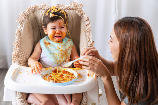 Unhappy Toddler Child With Tomato Sauce On Her Face Crying With Her Mother Sitting Together Beside In The Room. Upset Messy Little Girl Mix Race Hungry Eating Spaghetti With Mom At Home.