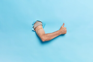 Horizontal shot of unrecognizable human breaks arm through paper blue background keeps thumb up recommends vaccination against coronavirus blue wall. Mass immunization and health care concept