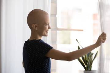 Happy millennial bald woman standing near window, opening curtains welcoming new day, feeling inspiration in fight with cancer, dreaming of healthy life, enjoying every day at home or oncology center.