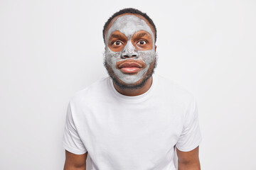 Portrait of surprised young African American man stares surprised at camera applies clay nourishing facial mask to remove fine lines and pores dressed in casual t shirt isolated over white background.
