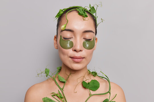 Asian Beauty Woman Cares About Skin Undergoes Facial And Body Treatments Keeps Eyes Closed Applies Patches Under Eyes Poses With Naked Body With Green Plant Around Isolated On Grey Studio Wall