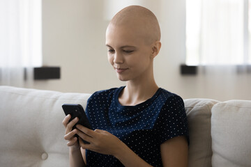 Relaxed young bald after chemotherapy woman sitting on couch, enjoying holding cellphone, web...