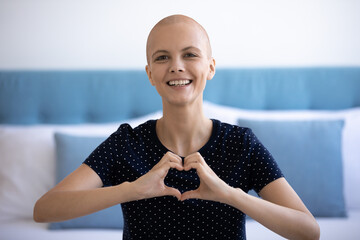 Happy young hairless woman making heart symbol with fingers, showing support to all people who...