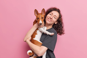 People and pets concept. Positive natural woman with curly dark hair carries her basenji dog spend...