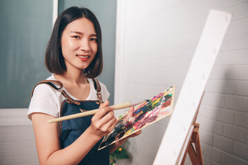 Asian woman artist painting picture with blank canvas side view, using paint brush and colour...