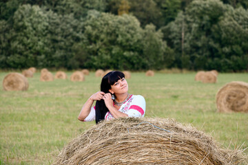 Naklejka na ściany i meble Ukrainian girl braids a braid against the background of a mown field. Standing behind a round haystack. Summer mowing.