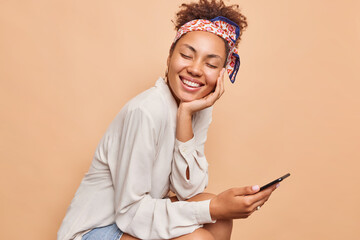 Horizontal shot of pretty millennial ethnic woman smiles happily keeps eyes closed browses mobile phone wears shirt and kerchief tied over head isolated over beige background. Technology concept