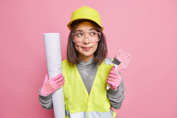 Pensive Asian female maintenance worker holds rolled blueprint painting brush looks away...