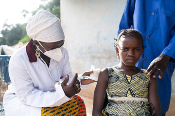 Worried little African girl undergoing influenza vaccination by a black nurse with protective face...