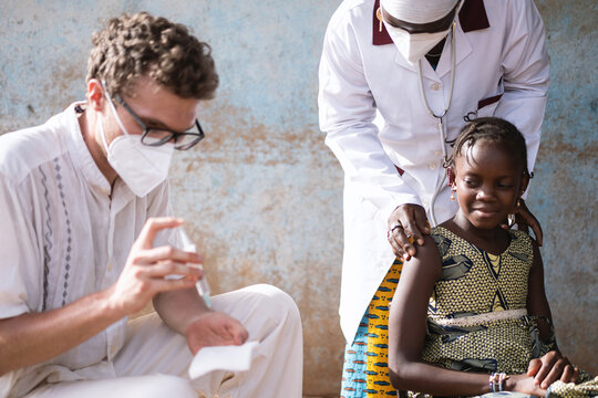 Male caucasian doctor preparing syringe, with little African girl waiting for her injection, reassured by a gentle black nurse bending over her