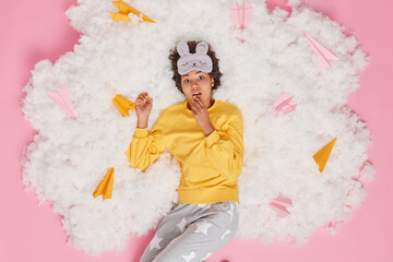 Overhead shot of surprised curly haired woman awakes in morning after seeing unexpected dream gazes shocked at camera keeps hand on chin wears nightwear poses on white cloud pink background.