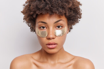 Beauty day. Serious curly haired woman with healthy skin applies silver patches under eyes looks directly at camera stands topless against white background. Wellness rejuvenation wellbeing concept