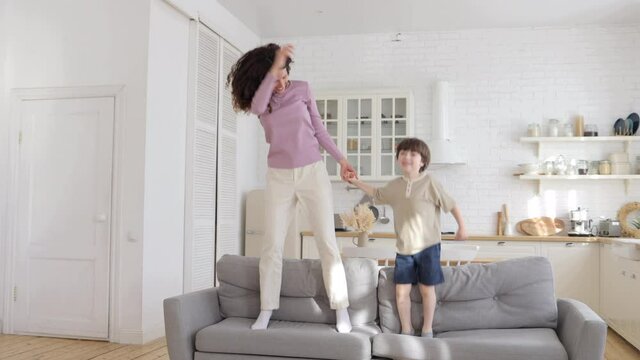 Crazy energetic family of two: mom and son active dance on couch in living room. Preschool boy funny activity indoors with young happy mother, sister or baby sitter. Carefree parent and child bonding